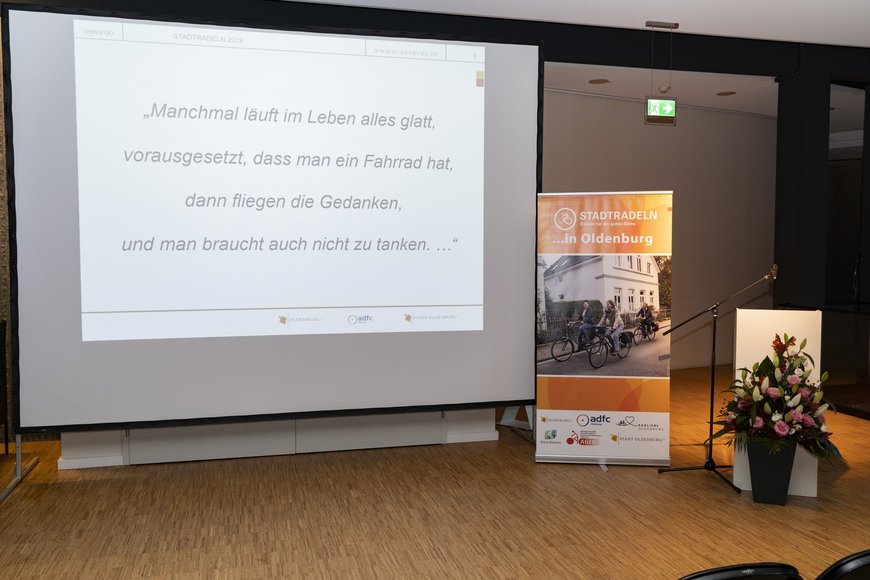 Start der Abschlussveranstaltung mit einer Präsentation auf einer Leinwand. Dabei ist eine Zeile aus dem Lied von Max Raabe „Manchmal läuft im Leben alles glatt, vorausgesetzt, dass man ein Fahrrad hat, dann fliegen die Gedanken,und man braucht auch nicht zu tanken. …“ zu sehen, der als Fahrradfreundlichste Persönlichkeit 2019 das STADTRADELN prominent unterstützt hat. Foto: Stephan Walzl