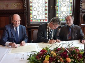 Bruce McDonald, Gerd Schwandner und Derek Osbourne sitzen an einem Tisch, im Vordergrund ein Blumengesteck, und unterzeichnen die Partnerschaftsurkunde. Foto: Stadt Oldenburg