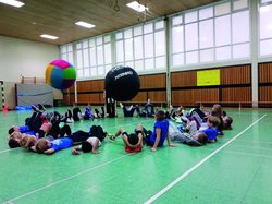 Zu sehen sind ungefähr zwanzig Kinder in einer Turnhalle. Sie liegen im Kreis auf dem Rücken und treten mit den Füßen in die Mitte, um zwei große Bälle hin und her zu bewegen. Foto: SSB e.V.