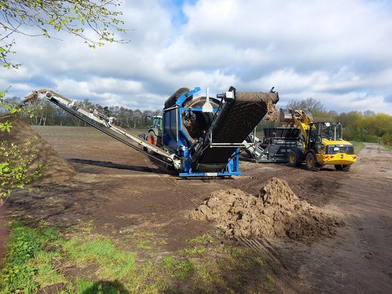 Die Säcke werden zunächst mit dem Bagger in den Schredder befördert, anschließend wird das Material durch die blaue Siebtrommel getrennt. Am Ende entstehen Haufen mit Sackmaterial aus Jute (vorne) beziehungsweise Sand (links). Foto: Stadt Oldenburg Die Säcke werden zunächst mit dem Bagger in den Schredder befördert, anschließend wird das Material durch die blaue Siebtrommel getrennt. Am Ende entstehen Haufen mit Sackmaterial aus Jute (vorne) beziehungsweise Sand (links). Foto: Stadt Oldenburg