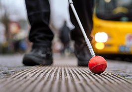 Beine und ein Blindenstock an einer Bushaltestelle mit Bus im Hintergrund. Foto: Izabela Mittwollen