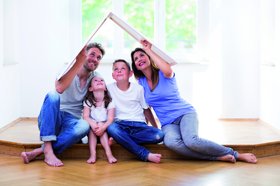 Auf dem Bild sitzt eine Familie mit zwei Kindern auf dem Boden und bildet mit zwei Plakaten über ihren Köpfen ein Dach als Haus.