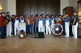 Die Shanty-Chöre aus Oldenburg und Rügen, Männer in weißen Latzhosen mit Gitarren und Akkordeons. Im Vordergrund ein Steuerrad eines Schiffes. Foto: Stadt Oldenburg