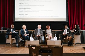 Podiumsdiskussion: Erastus Mwencha (2. von rechts) und Darby Gounden (1. von rechts) sitzen mit anderen Teilnehmenden im Stuhlkreis auf einer Bühne. Im Hintergrund ist eine Leinwand zu sehen. Foto: Patricia Kühfuss